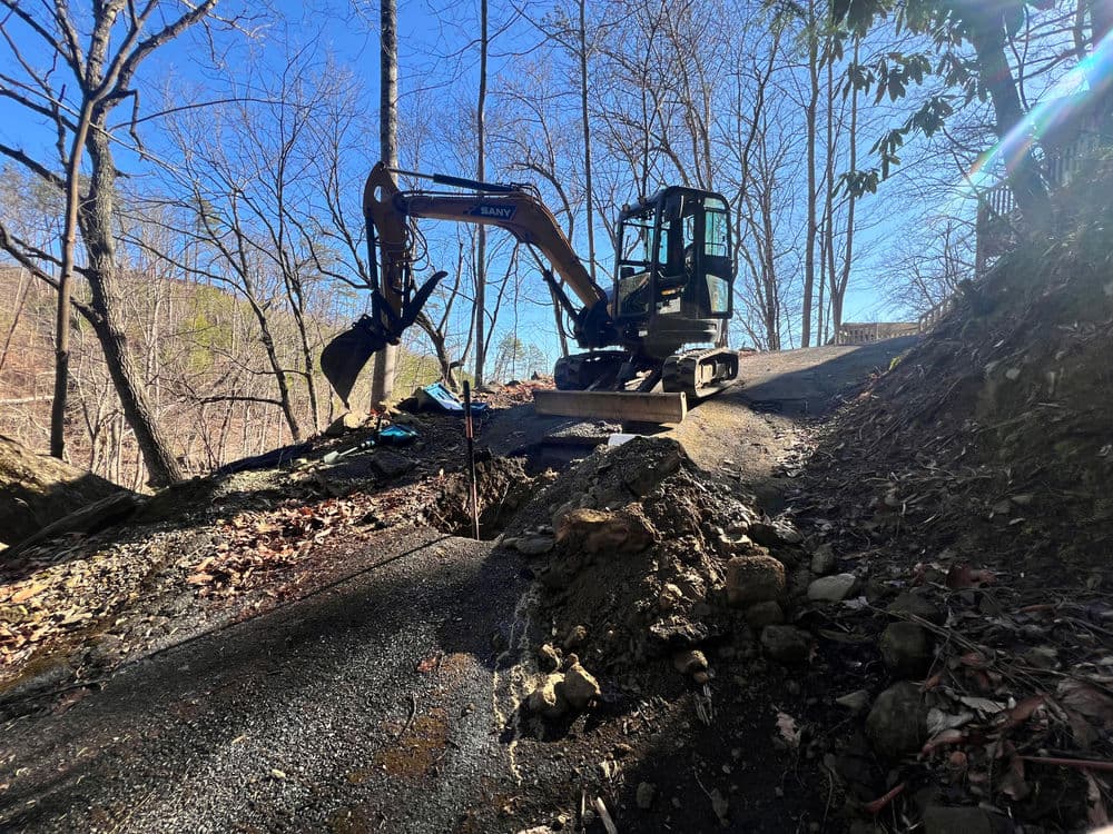 Excavator on a gravel road in a wooded area, surrounded by bare trees and blue sky.