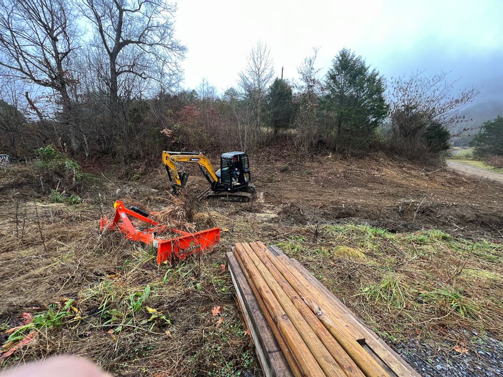 Backhoe excavator working on land clearing with fallen logs and equipment in a rural setting.