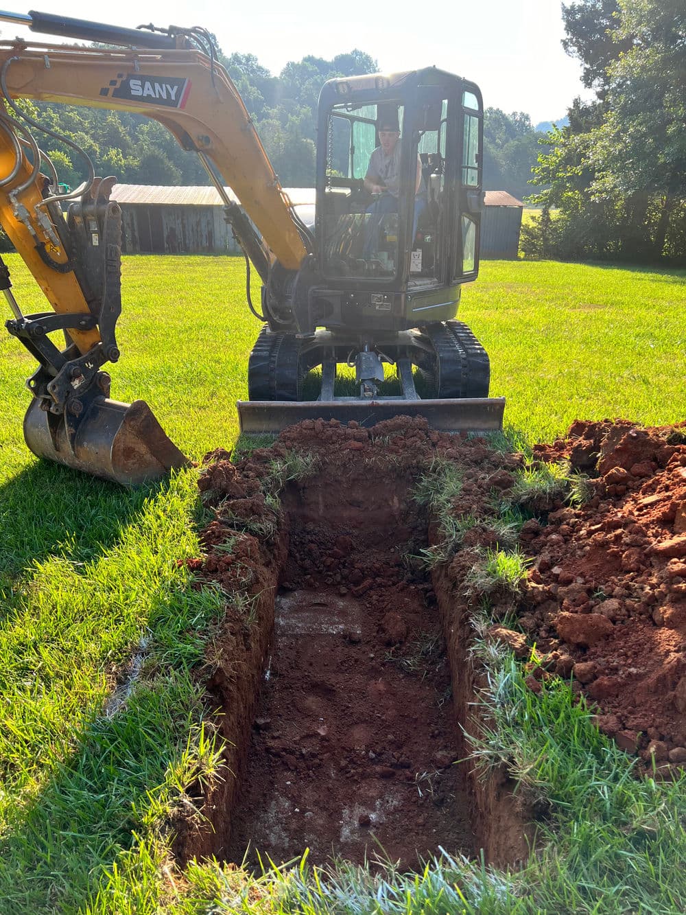 Excavator digging a trench in a grassy field with a driver inside.