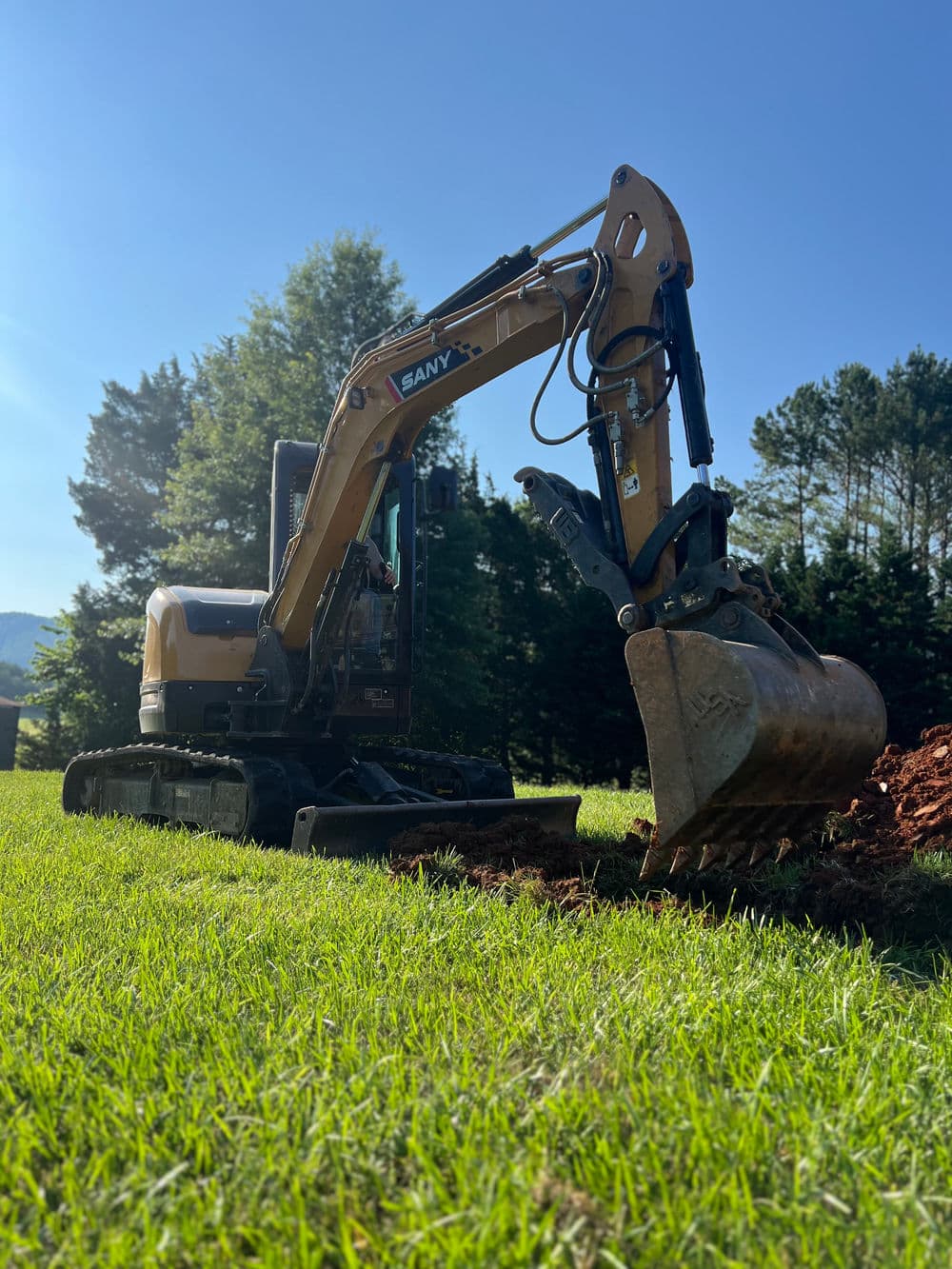 Excavator digging in a grassy field with trees and blue sky in the background.