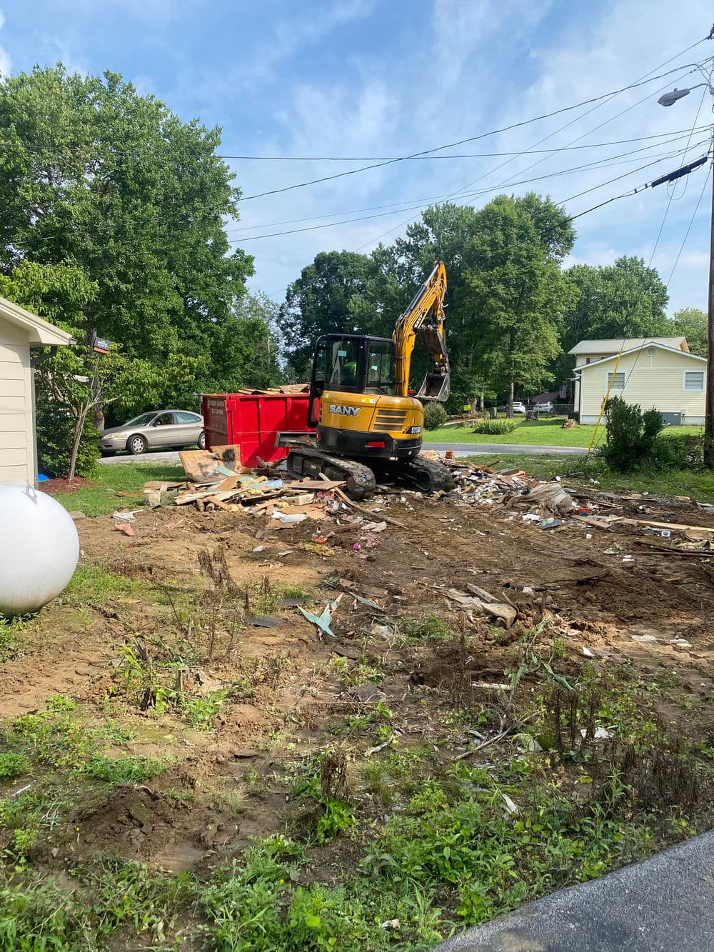 Excavator clearing debris and trash from a residential yard under a blue sky.
