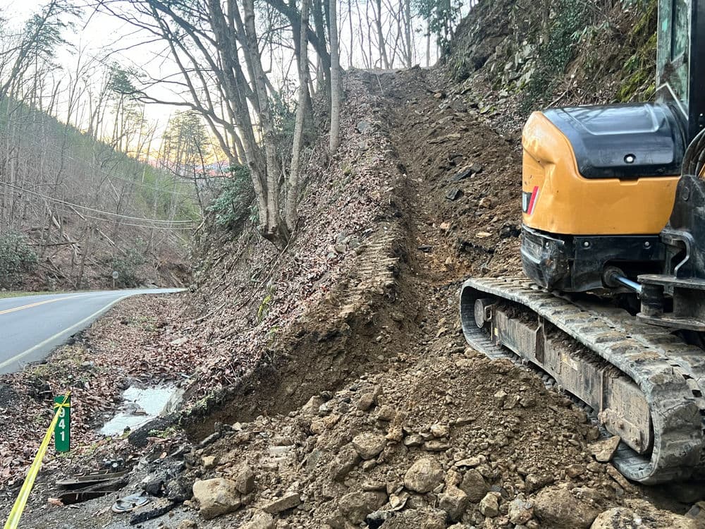 Excavator on a hillside preparing for road work, surrounded by trees and loose soil.