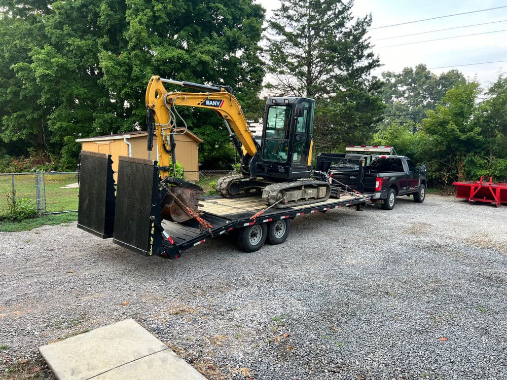 Excavator on flatbed trailer hitched to truck, parked in gravel area with trees in background.