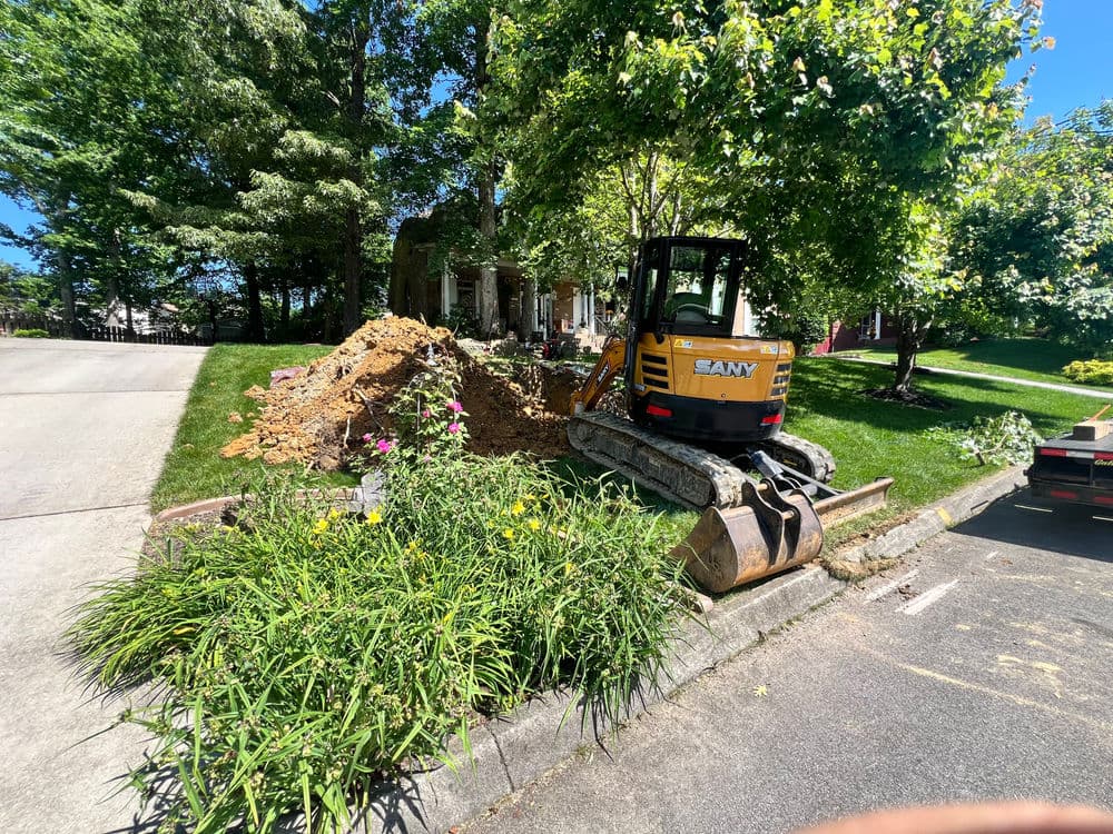 Excavator on a residential lawn next to a pile of dirt and flowers in the foreground.