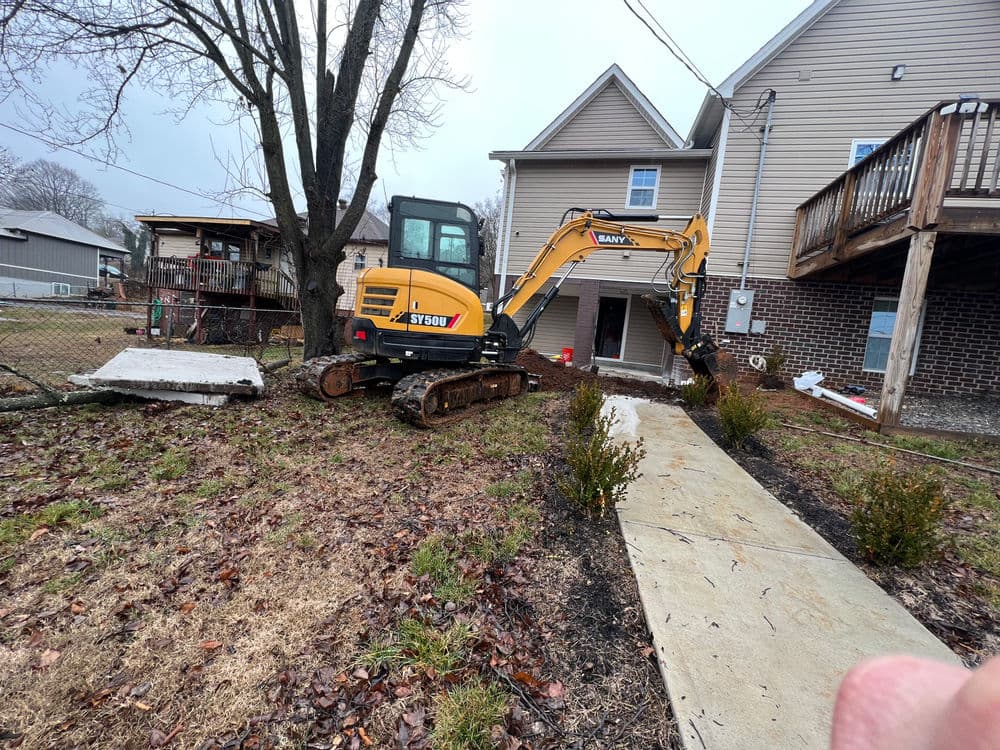 Excavator parked near a house, surrounded by wet ground and construction materials.
