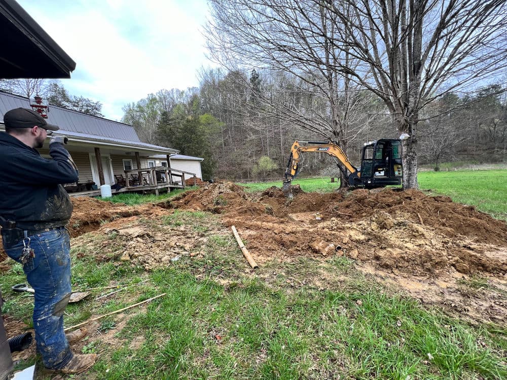 Excavator digging in yard while worker supervises near house and tree in rural setting.