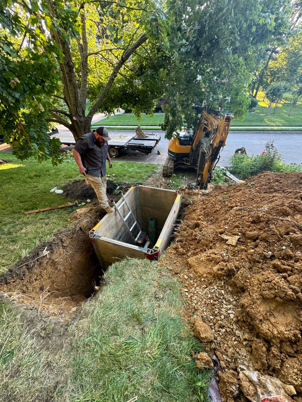 Construction worker digging a trench for a concrete vault with machinery and tools nearby.