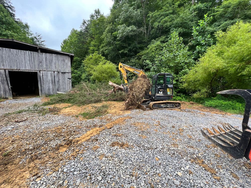 Excavator clearing debris near a barn in a wooded area. Gravel surface, cloudy sky.