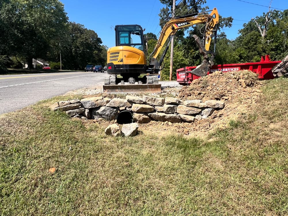 Excavator working on a drainage project with rocks and equipment near a road.