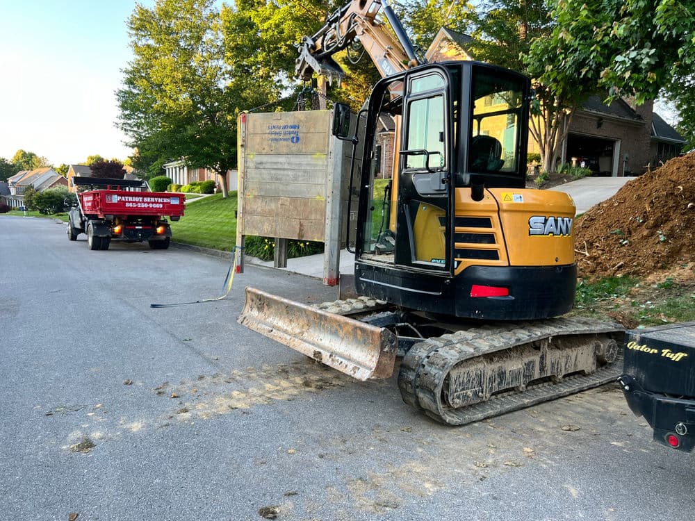 Excavator on residential street alongside a dump truck and construction materials.
