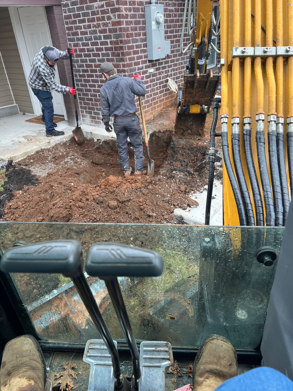 Men excavating a trench near a house, with construction equipment in the foreground.